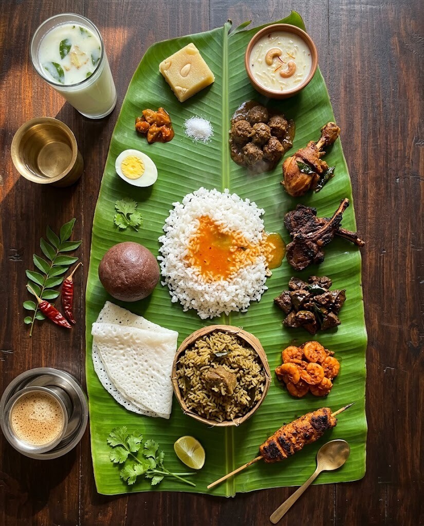 A full Karnataka non-veg spread laid on a banana leaf — rice, naati chicken, mutton kheema, neer dosa, ragi mudde, halwa and more, on dark wood with curry leaves and dried chilies